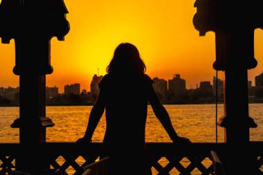 Genç bir kadın, Nil nehrindeki turuncu günbatımına bakıyor. Güneş arka planda Kahire şehrinin binalarında saklanıyor. Afrika