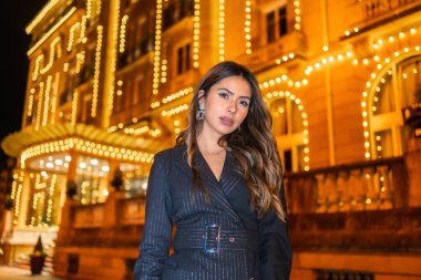 Noel çatışması, Kafkasyalı esmer kız gece şehirde Noel ışıklarının yanında gülümsüyor. Zarif siyah Noel ya da siyah çizmeli yeni yıl elbisesi