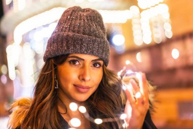 Yılbaşı ışıklarıyla Noel çekimleri, genç beyaz bir kız gece elinde güzel ışıklarla şehirde. Kış şapkalı, ışıklara bakan, şehvetli bakışlı.