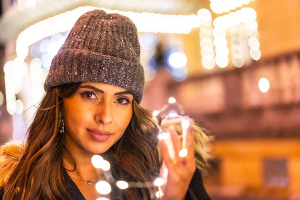 Yılbaşı ışıklarıyla Noel çekimleri, genç beyaz bir kız gece elinde güzel ışıklarla şehirde. Kış şapkalı, ışıklara bakan, şehvetli bakışlı.