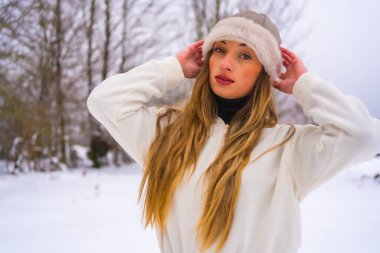 Kış yaşam tarzı, beyaz kışlık giysili beyaz Kafkas sarışının portresi ve karlı çam ağaçlarının yanında yünlü şapka, doğada tatil.