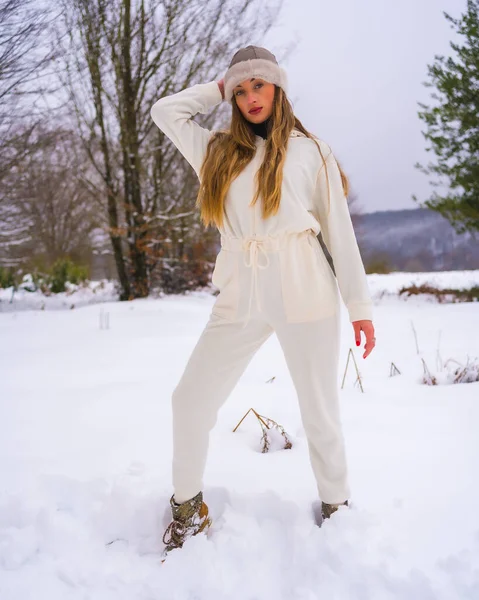 冬天的生活方式 金发碧眼的年轻姑娘 穿着白色的冬装 头戴羊毛帽子 站在雪白的松树旁 在大自然中度假 — 图库照片