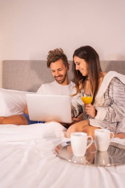 Kahvaltıda bilgisayara bakan pijamalı bir çift otel yatağında kahve ve portakal suyu, birbirine aşık bir çiftin yaşam tarzı..