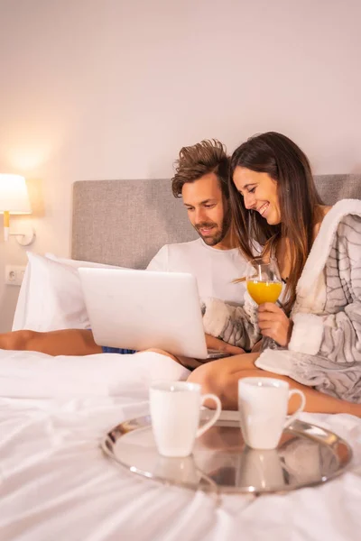 一对穿着睡衣的情侣 在早餐时看着电脑 在宾馆的床上看着橙汁 在恋爱中的情侣的生活方式 — 图库照片
