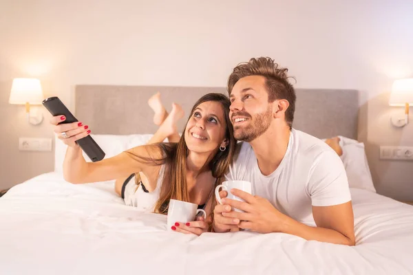 A couple in love watching a movie on TV on top of the hotel bed in pajamas, lifestyle of a couple in love.