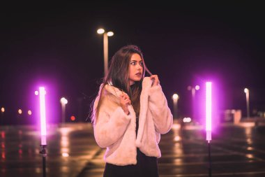 Pembe ışıkla aydınlatılmış boş bir otoparkta yün ceketli beyaz tenli bir genç. Şehirde gece şehir kış sezonu