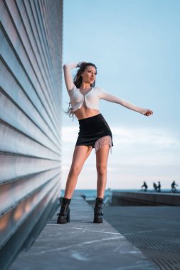 Şehirde mavi saatte, gri bir binanın yanında siyah çizme ve kısa siyah etek giyen güzel bir esmer kız resmi.