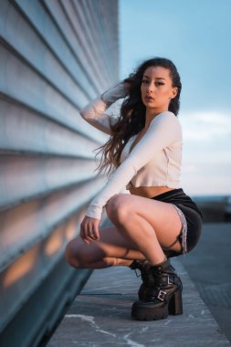 Siyah çizmeleri ve siyah kısa eteği olan, gri bir binanın yanında mavi saatte çömelmiş esmer bir Arap kızı poz vermek.