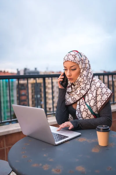 Arab girl in a white veil at the computer on the terrace of a coffe shop, making a video call. Young modern arab with new technologies, vertical photo