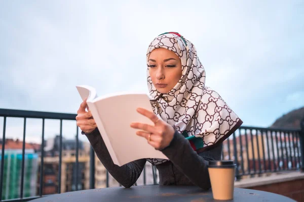 Arab girl with white veil at the computer on the terrace of a coffe shop, reading a beautiful book