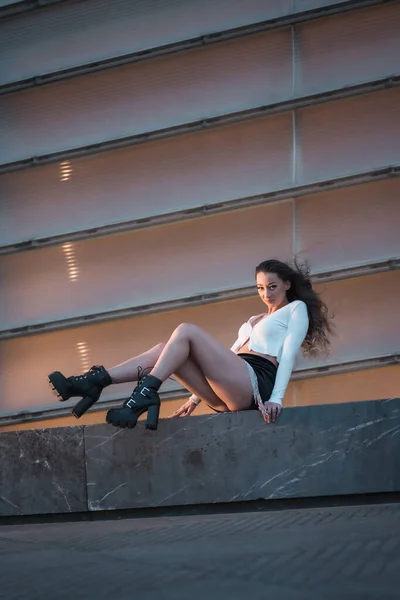 生活方式 一个漂亮的黑发女孩坐在一座灰色建筑旁边的蓝色时辰 穿着黑色靴子和黑色短裙 — 图库照片