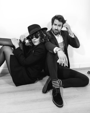 Siyah beyaz bir moda çekiminde yerde oturan beyaz bir çift şık takımlar ve güneş gözlüğü takıyor.