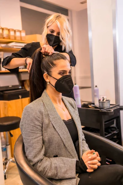 Caucasian hairdresser with mask finishing the hairstyle to the brunette client. Security measures of Hairdressers in the Covid-19 pandemic
