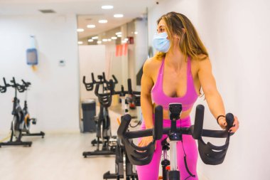 Koronavirüs salgını. Yüz maskesi takan sabit bisiklet eğitimi alan bir kız. Kapasitesi düşük spor salonları, sosyal mesafesi düşük ve yeni bir normal.