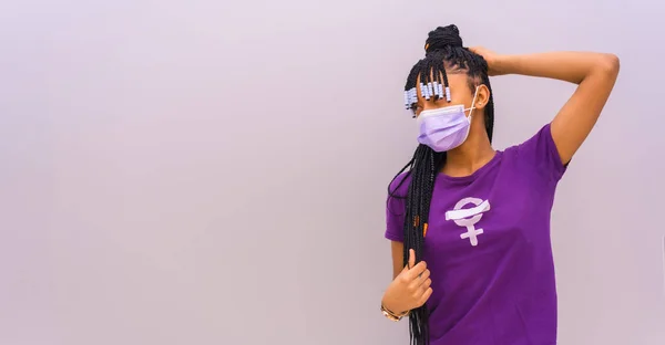 International women\'s day in the year of the coronavirus pandemic, covid-19. Dominican woman with feminist purple shirt and face mask, gray plain background with copy and paste space
