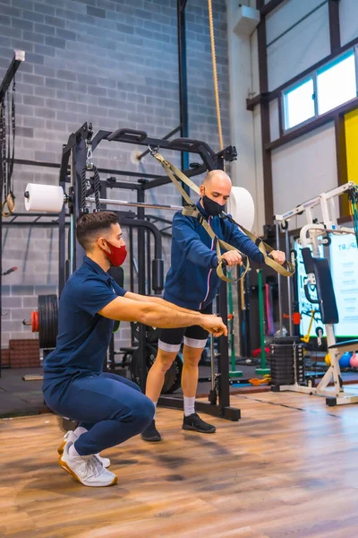 An athlete with an instructor in the gym performing exercises with strips in the coronavirus pandemic, a new normal. With protective face mask