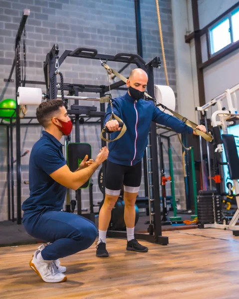 An athlete with an instructor in the gym performing exercises with strips in the coronavirus pandemic, a new normal. With protective face mask