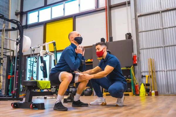 Personal trainer in the gym correcting the athlete\'s squats in the coronavirus pandemic, a new normal. With protective face mask