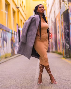 Bir şehir caddesinde krem renkli bir elbise giymiş siyahi Afrikalı bir kızla son moda şehir tarzı. Omzunda bir ceketle