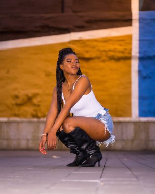 Şehirdeki bir sokakta örgü ören Afrikalı bir kızla şık bir şehir tarzı. Renkli arkaplanda beyaz gömlek ve şort