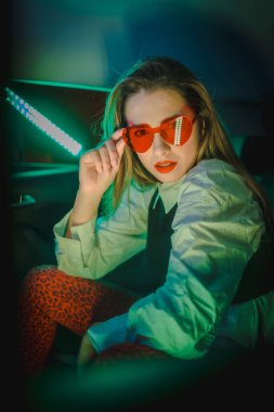 Kalp gözlüklü, sarışın, beyaz bir kadının arabasının arkasında yeşil neonlar olan şehir fotoğrafçılığı.