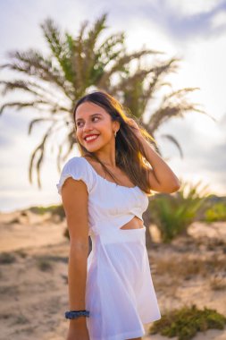 Beyaz elbiseli, dikey fotoğraflı bir palmiye ağacının yanında plaj tatilinden zevk alan genç beyaz bir esmerin yaşam tarzı.