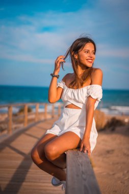 Yazın beyaz elbisesiyle sahilde tatilde olan genç esmer bir beyazın yaşam tarzı. Tahta kaldırımda oturuyor.