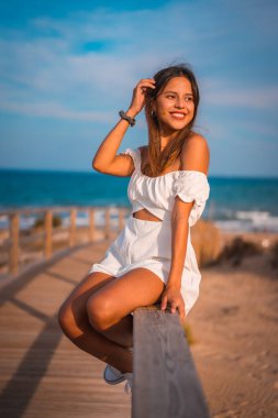 Yazın beyaz elbisesiyle sahilde tatilde olan genç esmer bir beyazın yaşam tarzı. Tahta kaldırımda oturuyor.