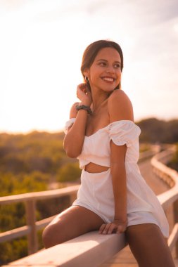 Yazın beyaz elbiseli, kumsalda tatilde olan esmer bir beyaz kadın, ahşap geçitte eğleniyor.