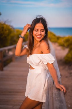 Yazın beyaz bir elbiseyle sahilde tatilde olan esmer bir beyaz, tahta yolda gülümsüyor.
