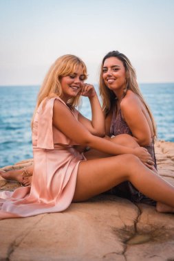 Bir çift beyaz kız çok güzel elbiseler içinde oturmuş deniz kenarında yazın tadını çıkarıyorlar.