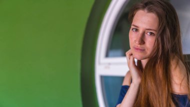 Genç, kızıl saçlı, mavi elbiseli beyaz bir ev satışının yanında gülümseyen beyaz bir beyaz kadın.