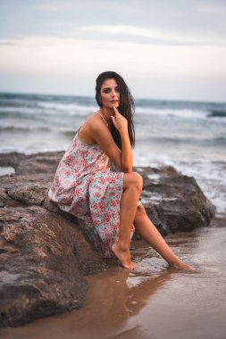 Beyaz kumaştan elbisesi ve çiçekleri olan esmer bir beyazın fotoğrafı. Denizde, sahilde tatilin tadını çıkarıyor.