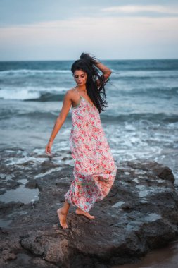 Kumsalda deniz kenarında tatilin tadını çıkaran, beyaz kumaşlı, çiçekli beyaz bir beyaz kadın pozu.