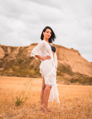 Beyaz bir elbise ve siyah bir bikini giyen esmer bir mankenin portresi güzel bir çölde, bir yaz öğleden sonra, dikey bir fotoğraf.