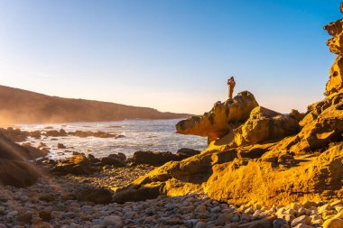 San Sebastian yakınlarındaki Pasajes kasabasındaki Jaizkibel Dağı 'ndaki taş koyda gün batımında fotoğraf çeken genç bir kadın. Bask Ülkesi