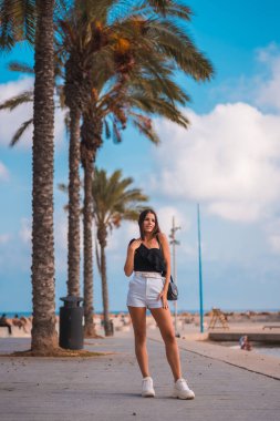 Yaz yaşam tarzı, beyaz şortlu beyaz esmer ve Akdeniz kıyısında palmiye ağaçlarının yanında yürüyen siyah tişörtlü.