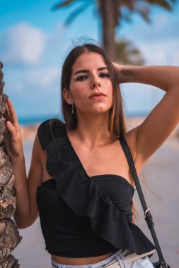 Akdeniz kıyısında beyaz şortlu ve siyah tişörtlü, palmiye ağaçlarının yanında esmer bir beyazın gülümsemesi.