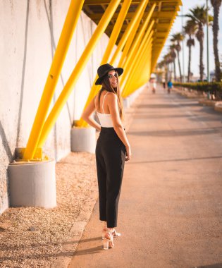 Genç bir esmer, topuklu, pantolonlu ve siyah şapkalı.