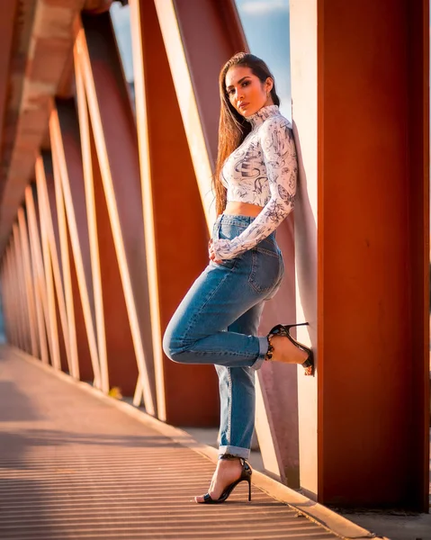 日落时在城里一个穿着牛仔裤的黑发拉丁姑娘 坐在橙色的桥旁边 — 图库照片
