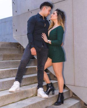 Mavi takım elbiseli ve yeşil elbiseli beyaz bir çiftin şık duruşu, şehrin merdivenlerinde birbirlerine sarılmaları.