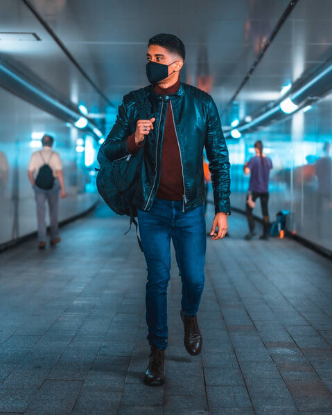 Fashion lifestyle, portrait of a young Latino in the city subway. Jeans, leather jacket and brown shoes. In a pandemic with a mask