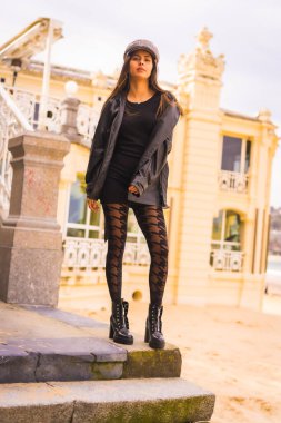 Yaşam tarzı, kısa siyah bir elbiseyle merdivenlerde oturan beyaz esmer, plajın yanında bereli ve ekose çoraplı, dikey bir fotoğraf.