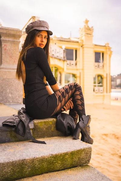 Yaşam tarzı, beyaz esmer, kısa siyah bir elbiseyle merdivenlerde oturuyor, bereli ve ekose çoraplı, yaz tatilinde bir plajın yanında.