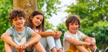 Lifestyle of three brothers smiling in a park sitting next to a tree, friends and brothers enjoying as a family