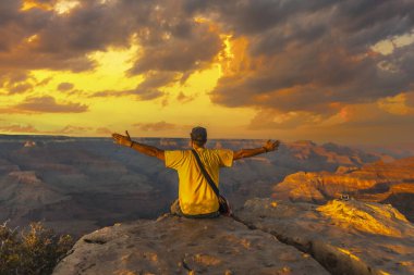 Büyük Kanyon Powell Point 'te Sunset' te elleri açık oturan ve sarı bir gömlek giyen genç bir adam. Arizona, dikey fotoğraf.