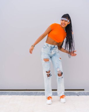 Örgülü genç bir tuzak dansçısı. Turuncu tişörtlü, gri arka planda kovboy pantolonlu, örgü ören ve gülümseyen Afrikalı etnik grubun zenci kızı.