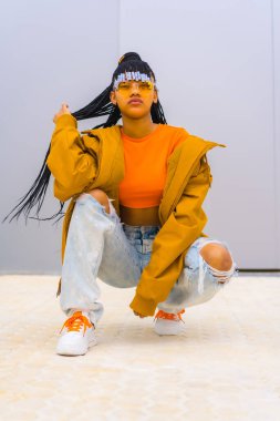 Örgülü genç bir tuzak dansçısı. Tişörtlü, turuncu güneş gözlüklü ve ceketli siyahi etnik kız baştan çıkarıcı yüzüyle kameraya bakarak çömeldi.