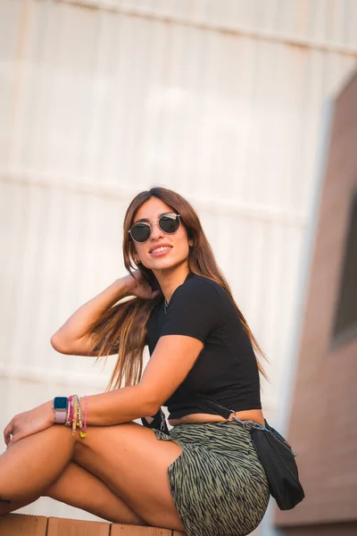 一个年轻迷人的高加索黑发姑娘 戴着太阳镜 坐在城市里 欣赏着白墙背景的阳台 她的街头风格很迷人 — 图库照片