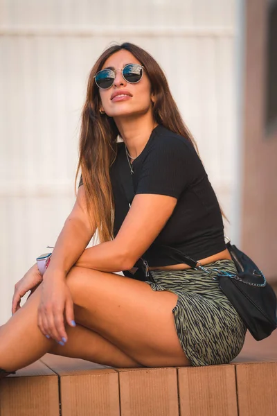 一个年轻迷人的高加索黑发姑娘 戴着太阳镜坐在城市里 欣赏着白墙背景的天鹅绒 垂直的照片 街道风格 — 图库照片